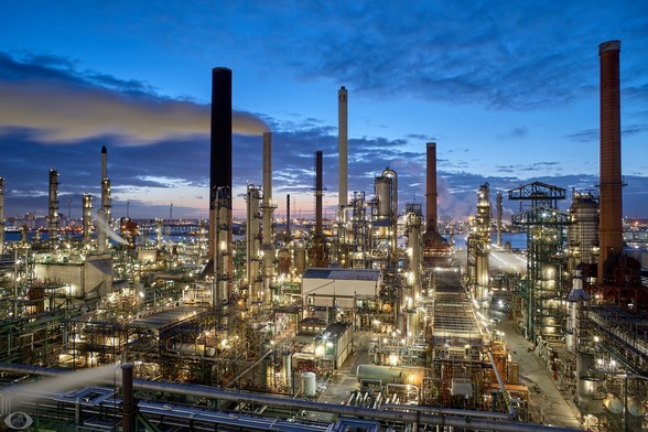 A bunch of chimneys, pipelines, reactor vessel, and other machinery in various dimensions and seemingly criss-cross placed and connected together, spewing smoke and steam in the blue hour and still lighted (providing a yellow-green gleam.) Behind it a river is barely visible. Barely visible, the sun is setting.