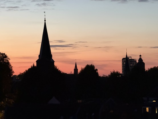 A fat church steeple is seen against the twilight skyline with the foliage of trees obscuring the main building, so that it looks like it's growing up out of some sort of mound covered with vegetation. No detail is visible in the foreground because of the darkness. Several other steeples and a hirise building are also visible against the sky. There are scattered small clouds.