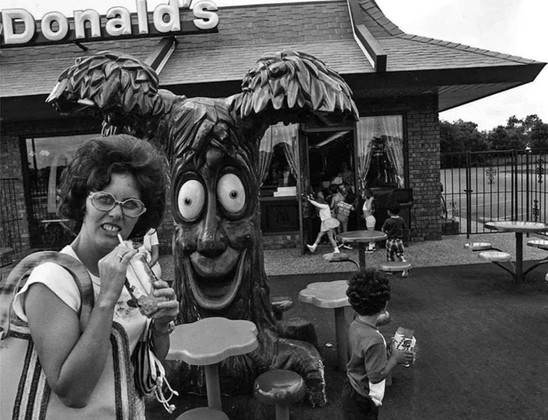 A woman with bad hair and eyewear sipping a drink in the McDonalds Play Area in the 70s or 80s.