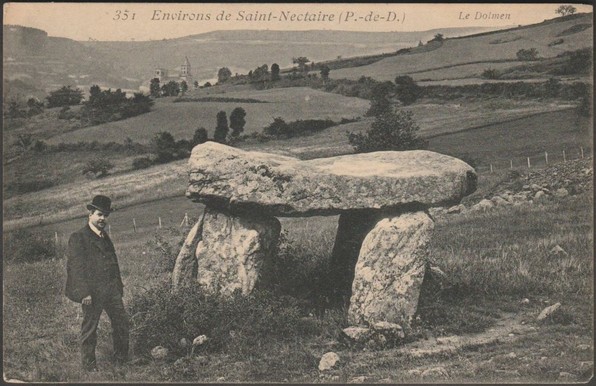 A printed vintage postcard showing a gentleman in bowler hat standing beside the Dolmen du Parc or Parc du Dolmen, close to Saint-Nectaire-le-Bas in Puy-de-Dôme, France. The 3.7m by 2.4m capstone sits on four supporting stones, with another lower stone alongside, all being volcanic stone from the area. The town of Saint-Nectaire can be seen in the valley below.