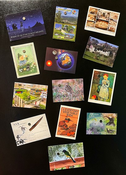 Thirteen different postcards received from around the world over the past year, displayed at jaunty angles with magnets on a black refrigerator door.