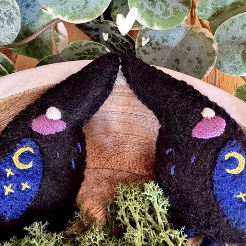 Picture of two handmade crow plushies made with black felt for the body, and dark blue for the wings. They are facing one another on a wooden background, surrounded with leaves and moss, their beaks touching. Three white hearts are doodled above their beaks. 
They both have round white eyes and a big pink blush embroidered on their faces, as well as 3 small blue line on their chest. On their wings, three stars and a sickle moon are embroidered in yellow on their wings.