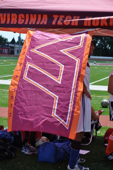 A Virginia Tech flag in maroon and orange is prominently displayed, partially obscured by a person. The background shows a sports field, and belongings like bags and a soccer ball are visible on the ground.