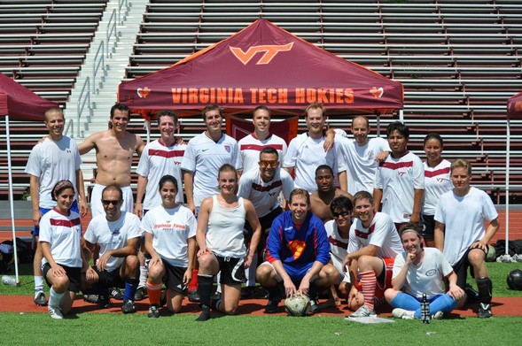 A group photo of a coed soccer team poses on a field. They are wearing various soccer jerseys, with some in Virginia Tech colors. A few members are kneeling in front, while others stand behind them, with a Virginia Tech tent visible.