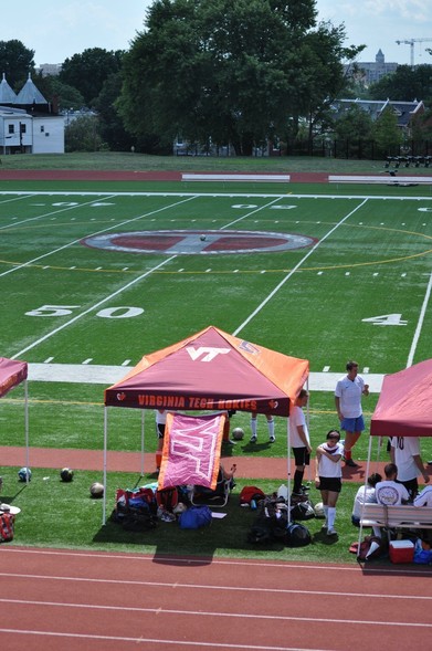 A sports field featuring a Virginia Tech tent and various soccer players. Soccer equipment and bags are visible near the tent, with a discernible field marking at the center. The background includes trees and a few buildings.
