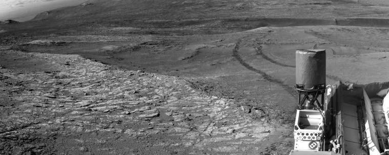 A black and white panorama capturing a Martian landscape, with the rear section of the Curiosity rover in the lower right corner. Rover tracks are receding away in the distance.