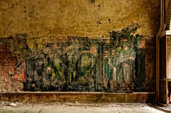 marode Ziegelwand mit nur noch zum kleinen Teil erhaltenen Wandmalerei aus der Nachkriegszeit. Zu sehen sind noch Personen in Einheitskleidung