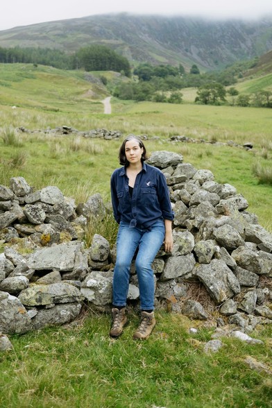 A woman in a navy work shirt and jeans sits on a partially built drystone wall in a green Scottish glen. Behind her, rolling hills and a mist-covered mountain rise in the distance, with a winding path cutting through the landscape. The scene is lush and overcast, evoking a quiet, grounded atmosphere.