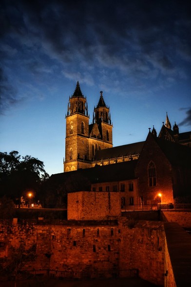 A captivating low-angle night shot captures the majestic Magdeburg Cathedral, illuminated in warm golden light against a deep twilight sky. Its two prominent spires dominate the upper frame, showcasing intricate architectural details. Below the cathedral, ancient, rough-hewn stone walls and buildings are visible, also bathed in the warm glow of streetlights, leading down to a lower, darker foreground. Silhouetted trees and subtle cloud formations in the blue sky add to the dramatic ambience of this historic German landmark.
