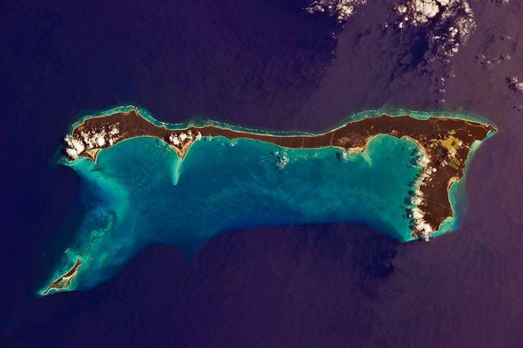 an aerial photograph of a thin island, it looks like a hammer, light aqua water around it, around it is purple water, some small buildings on it, this island was san salvador but was renamed