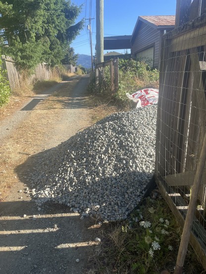 The image depicts a narrow, unpaved road flanked by wooden fences on both sides. The road is bordered by dry grass and sparse vegetation, with a large pile of gray gravel prominently placed on the right side, partially obstructing the path. A red and white bag is visible near the gravel pile, suggesting recent construction or landscaping activity. In the background, a house with a red roof is partially visible, along with a utility pole and power lines. The sky is clear and blue, indicating a sunny day. The scene is set in a rural or suburban area, with mountains visible in the distance.