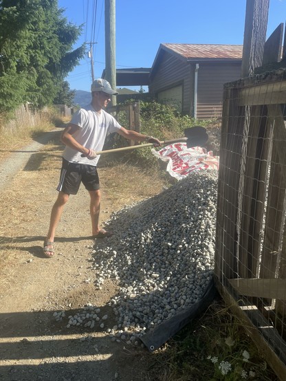 The image shows a person engaged in manual labor, specifically shoveling gravel. The individual is wearing a white t-shirt, black shorts with white stripes, a white cap, and sandals. They are using a shovel with a long wooden handle to transfer gravel from a bag onto a pile. The gravel is light gray and appears to be small, rounded stones. The person is standing on a dirt path next to a wire fence, with a brown house and a garage visible in the background. The sky is clear and blue, indicating a sunny day. The scene is set in a residential area with trees and power lines visible in the background