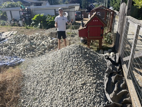 The image shows a young man standing in a backyard area. He is wearing a white t-shirt, black shorts, and a cap, and is holding a shovel. In the foreground, there is a large pile of gray gravel. To the right, there is a red chicken coop with a brown roof, situated next to a wire fence. The background includes a house with a white exterior, a car parked nearby, and some greenery. The ground is covered with gravel, and there is a black tarp on the ground to the right of the gravel pile. The scene appears to be a residential backyard with ongoing construction or landscaping work.
―