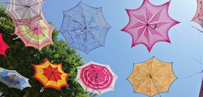 Bunt gemusterte  - gelb, pink, blau,weiss, grün - gehäkelte Schirme hängen unterm blauen Himmel und einem grünen Baum.