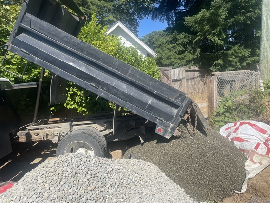 A small dump truck with a black box is seen mid-pour of pea gravel beside the drain rock just inside the fence, but not quite all of it as the truck is too big to turn into the yard