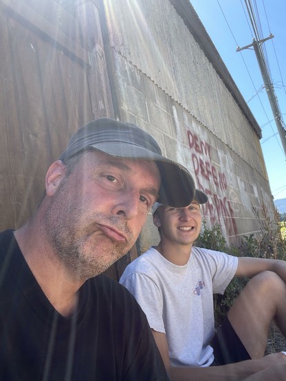 Two hot guys sitting down in the tiny sliver of shade beside the neighbours cinder block garage that has "Deny Defend Depose" written in red spray paint
