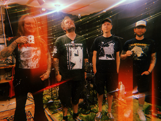 The four members of the band pose for the pic. They are dressed in black and look very cool and a bit smiley. One of them is holding a cup of coffee