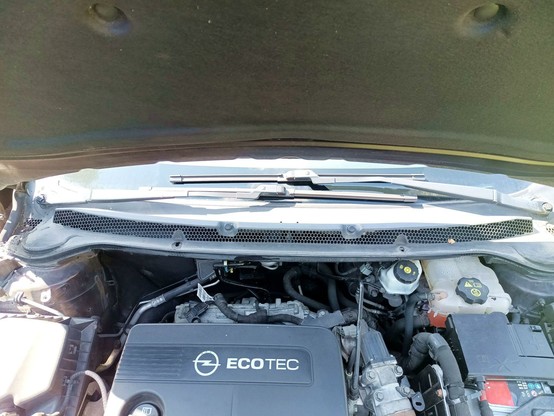 Under hood view of a car with the window wipers and ventilation drain box visible at the top, not opened yet, only the thick foam seal is pulled off.