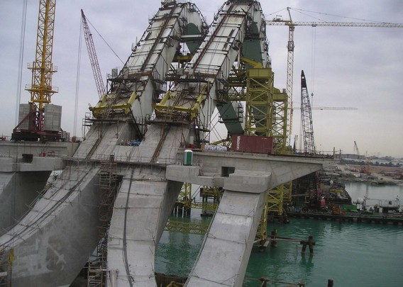a photograph of a large construct of steel and concrete, it curves upward, two large concrete strips, water underneath, many cranes around it