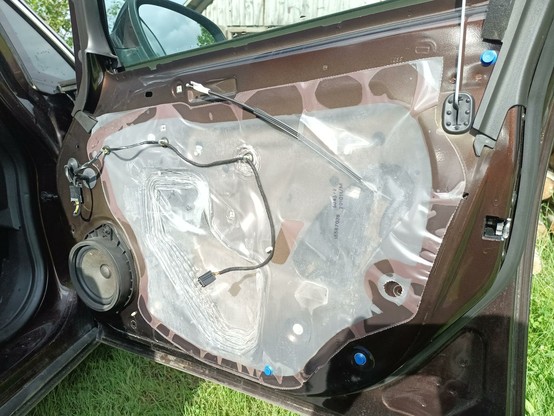 A car door with the panel removed. There's a speaker on the left. A large gap in the inner steel part is covered with a semi-transparent whiteish piece of thick plastic foil. It has many gaps all around where the adhesion of the plastic to the metal has failed and smal gaps have opened. Some of these had water droplets in them. Various plastic clips and cables and a bowden cable are routed in the door.