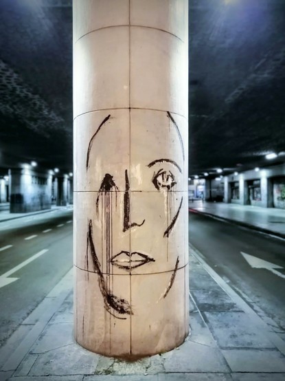 Sous un pont, au centre de l'image, une colonne beige sur lequel est dessiné, par quelques traits noirs, le visage d'une femme.
De part et d'autre de cette colonne, les rues.
Photo de nuit.