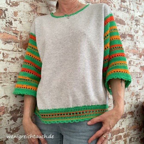 Eine Frau steht an einer Mauer, sie trägt ein helles Sweat Shirt mit bunten Häkelbordüren.