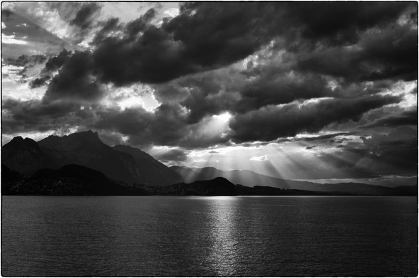 Sonnenstrahlen brechen durch Wolken über einem ruhigen See mit Bergen im Hintergrund