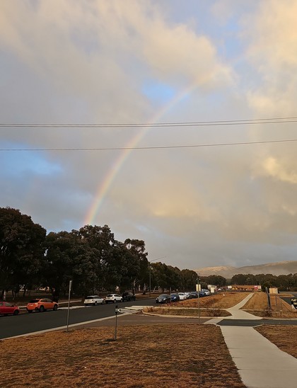 A bright rainbow is visible against a cloudy sky. The ground has a footpath running alongside a road where cars are parked, with a dry grass verge. Locals will recognise the Tuggeranong Archery Club at the end of the road.