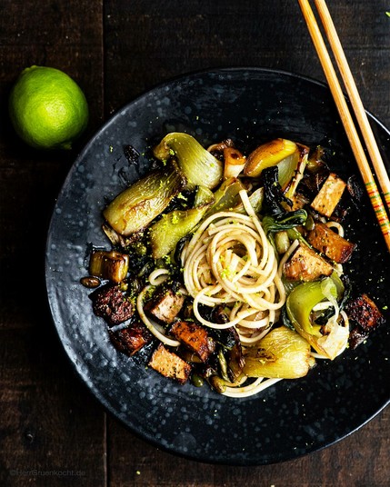 Dunkler Holzuntergrund. Darauf ein schwarzer, runder Pastateller. Darin: Udon-Nudeln mit Pak Choi, Räuchertofu, Edamame und geriebener Limette.