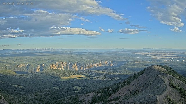 Beautiful colors and panoramic views from the Mount Washburn Fire Lookout camera last night. The foreground shows a section of the trail system used to access the mountain. Lit by the sun, the Grand Canyon of the Yellowstone cuts through the center of the image. In the distance on the right, the Tetons in Grand Teton National Park can just be seen. Blue sky, an arrangement of clouds, green vegetation, sunlit canyon walls, combine to create a wonderful scene from one of the most spectacular sites in Yellowstone National Park.