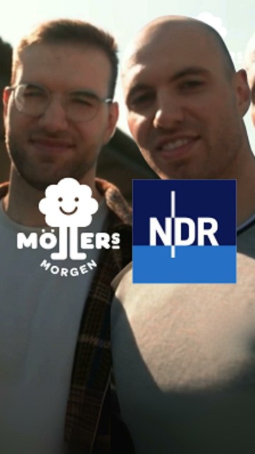 Das Bild zeigt zwei Personen, die sich für ein Selfie zusammen posieren. Die Person auf der linken Seite trägt eine Brille und ein hellgraues T-Shirt mit dem Logo "Möllers Morgen", während die Person auf der rechten Seite ein dunkles T-Shirt trägt. Beide Personen lächeln in die Kamera. Im Vordergrund des Bildes ist das Logo des NDR zu sehen, was darauf hinweist, dass das Bild möglicherweise im Zusammenhang mit einer Sendung oder einem Bericht des NDR steht.