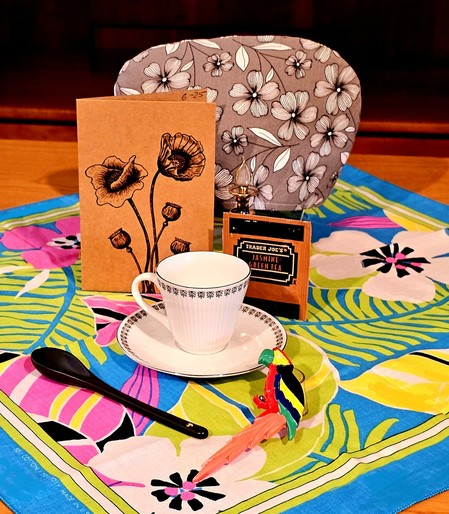 Tea tableau on a tropical tea towel in hot green, pink, and turquoise with black accents, a black and white floral cozy, a carved wooden parrot, white and black china and a woodcut card with an antherium design. The tea is Jasmine green tea from Trader Joe's