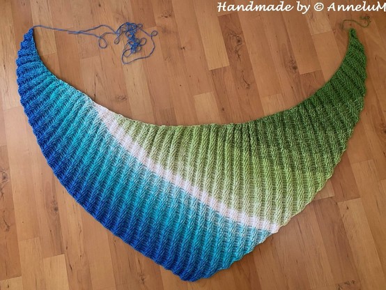 Das Bild zeigt ein gehäkeltes Dreiecktuch, das auf einem Laminatboden ausgebreitet liegt. Das Stück hat eine elegante, halbkreisförmige Form und zeigt einen sanften Farbverlauf von Blau über Türkis und Weiß bis hin zu Grün. Die Textur der Wolle weist ein rippiges Muster auf. Einige Fäden des restlichen Garns liegt unordentlich auf dem Boden. Im oberen rechten Bereich des Bildes ist der Text "Handmade by © AnneluM" zu sehen, was darauf hinweist, dass es sich um ein handgefertigtes Stück handelt.
Bereitgestellt von @altbot, privat und lokal generiert mit Ovis2-8B