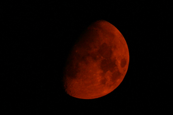 Photograph of a large, red waxing Moon, slightly more than half full with the visible part to the bottom right. Smoke particles in the sky have given the moon a very red tint.
