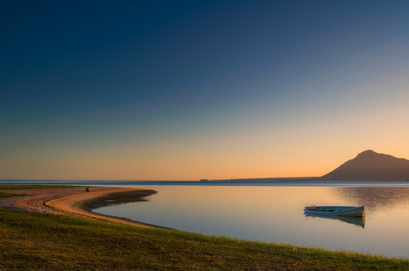 Sitzende Person am Ufer einer Lagune im Morgengrauen, ruhige Wasseroberfläche mit Boot, Berg im Hintergrund, entspannte Atmosphäre zum Stressabbau