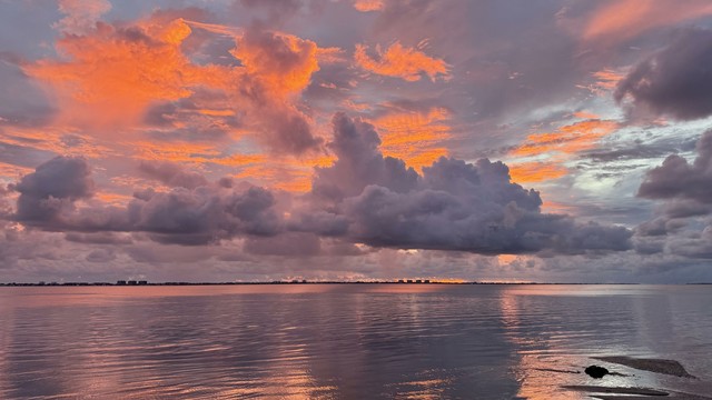 A scenic sunset over the bay. The sky is adorned with vibrant orange and pink clouds, reflecting on the water's surface, while a dark silhouette of land is visible in the distance.