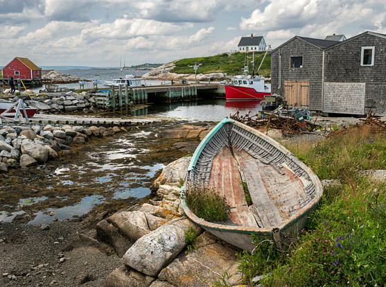 A small harbor at lowtide. An old rowboat sits up on land amidst small boulders and wild flowers. In the background a pier and a red fishing boat rest on the water. Clouds and blue sky up top.