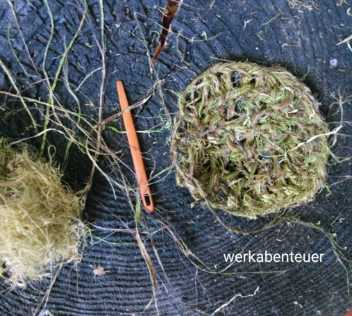Auf einem schwarzen Holzstück liegen grüne Fasern, eine Holznadel und ein angefangenes, fasergrünes, kreisförmiges Strickstück in einer Art Netzstruktur.
