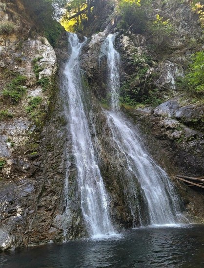 A cascading waterfall plunging into a dark, reflective pool at the base, surrounded by steep, rocky cliffs covered in patches of green vegetation and small trees.