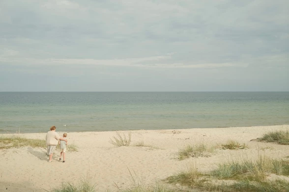 A woman and a child walk together along a sandy path towards the beach and the sea