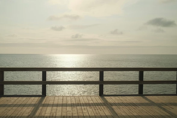 Sunlight reflects off the calm sea, viewed from a wooden pier railing