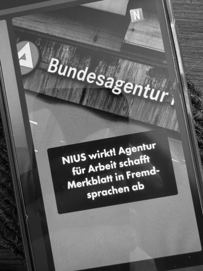 S/W-Foto der Webseite des Onlinemagazins _Nius_ am 13.8.2025, das Foto von Logo und Schriftzug der Bundesagentur für Arbeit, darunter eine Box mit dem Text "Nius wirkt! Agentur für Arbeit schafft Merkblatt in Fremdsprachen ab".