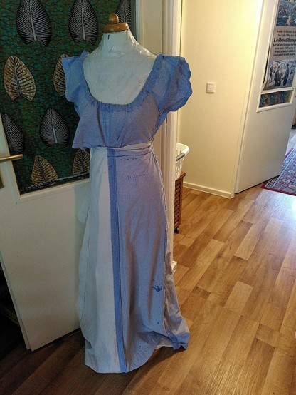 Picture of a homemade duct tape sewing dummy in a living room, on which a mockup made of old bedsheets embodies a floor length dress with a low neckline, a relatively high waist and short, fluttery sleeves. Only the left side of the bodice is pinned tidily into pleats that taper to the waist.