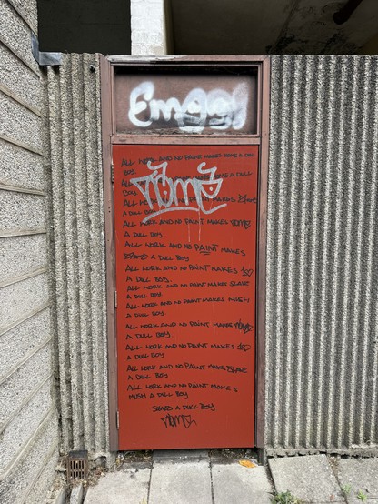 A weathered red door covered in graffiti, featuring repeated phrases like "All work and no paint makes YPM a dull boy" and various stylized signatures. The background displays a textured grey wall, and there's a drain on the ground.