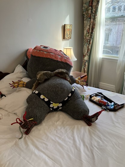 A large stuffed animal in a bikini that looks sort of like a labubu spread out on a hotel bed surrounded by the contents of the mini bar