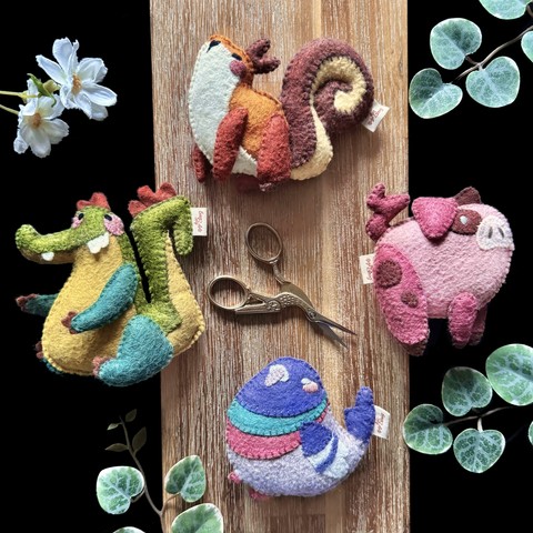 Picture of 4 handmade felt plushies on a black and wooden background, framed with decorative plants. 
There is a squirrel, a little pig, a very round pigeon, and a cheeky crocodile!