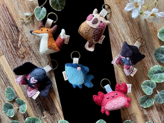 Picture of 6 handmade felt keychains photographed on a black and wooden background, framed with plants. 
The keychains are: a silly fox; a happy owl; a sweet whale; a happy crab; and two cute bats, one with its wings open, the other with its wings held closed thanks to small magnets concealed in the wings.