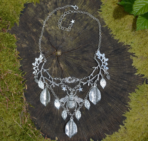 Large silver coloured bib necklace, lying flat on an old tree stump ringed with moss. A central bumble bee hangs from silver twigs, a v chain hangs underneath dotted with leaf pendants.