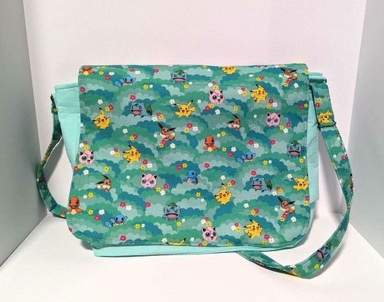 Messenger bag with Pokemon character print including Bulbasaur, Eevie, Pikachu, and others. Red, yellow and white flowers are scattered inbetween on a green and teal background.