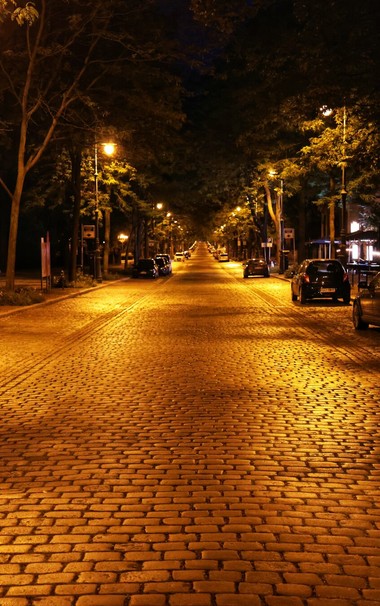 A night-time, low-angle photograph of a long, cobbled street lined with trees and streetlights on either side. The warm, yellow glow from the lamps illuminates the road, creating a strong sense of perspective as it stretches into the distance. Cars are parked along the kerbs on both sides of the street, and the bare branches of the trees are silhouetted against the dark sky.